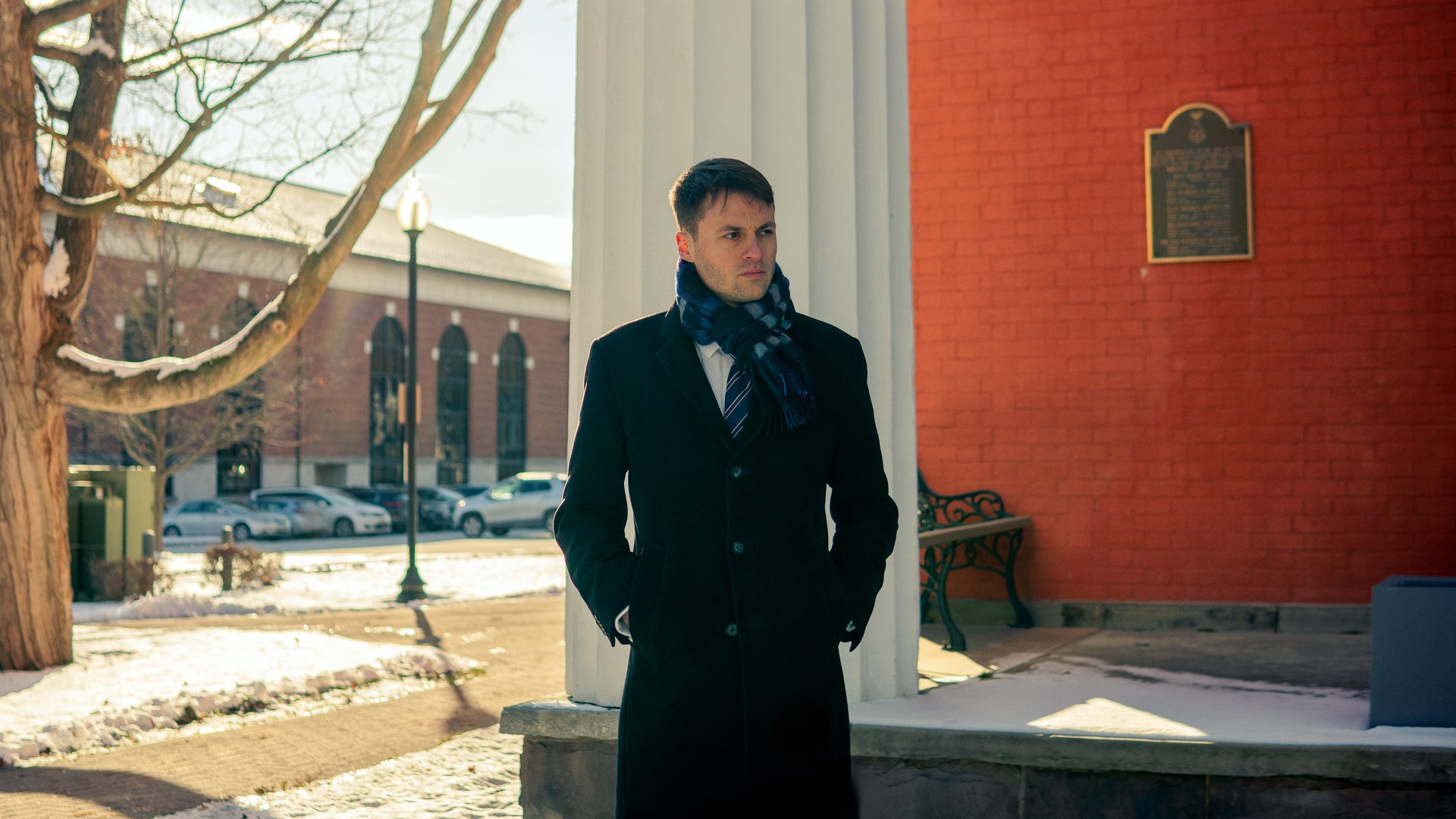 Thomas Plan in a dark winter coat and scarf, standing by a white column in Penn Yan and looking to the side.